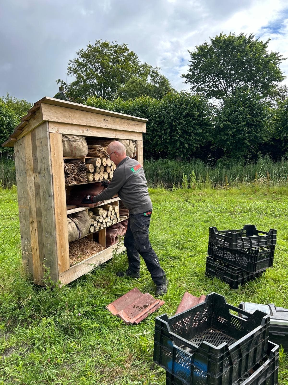 Een man in werkkleding van Van Beets vult een groot houten bijenhotel met vakken van riet, geboorde houtblokken, jute, stro en dakpannen, op een grasveld met bomen erachter.