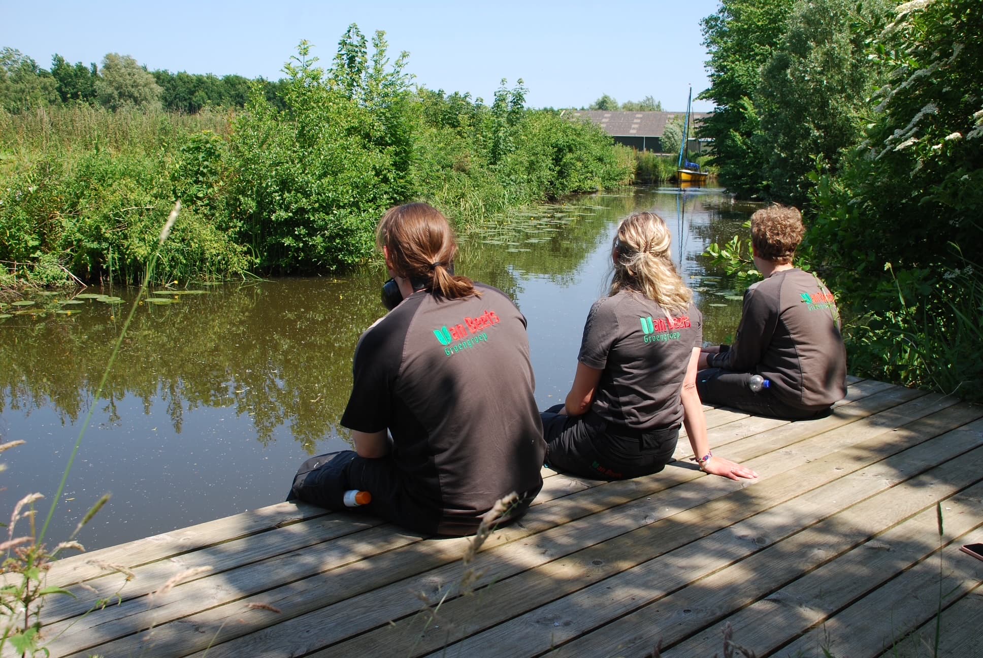 Drie collega's in werkshirts van Van Beets zitten op een houten steiger aan de vaart bij zorgboerderij Hiddema State, met riet, struiken en een zeilbootje in de verte