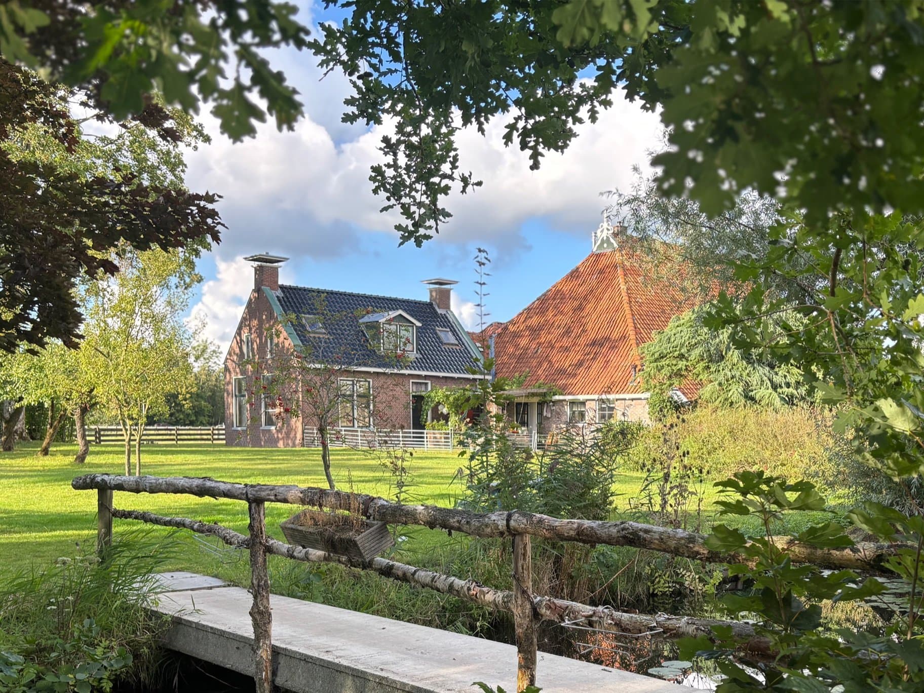 Het voorhuis en de schuur met oranje pannendak van zorgboerderij Hiddema State in Hempens, gezien tussen de bomen door, met een grasveld, een houten hekje en een bruggetje over de sloot.