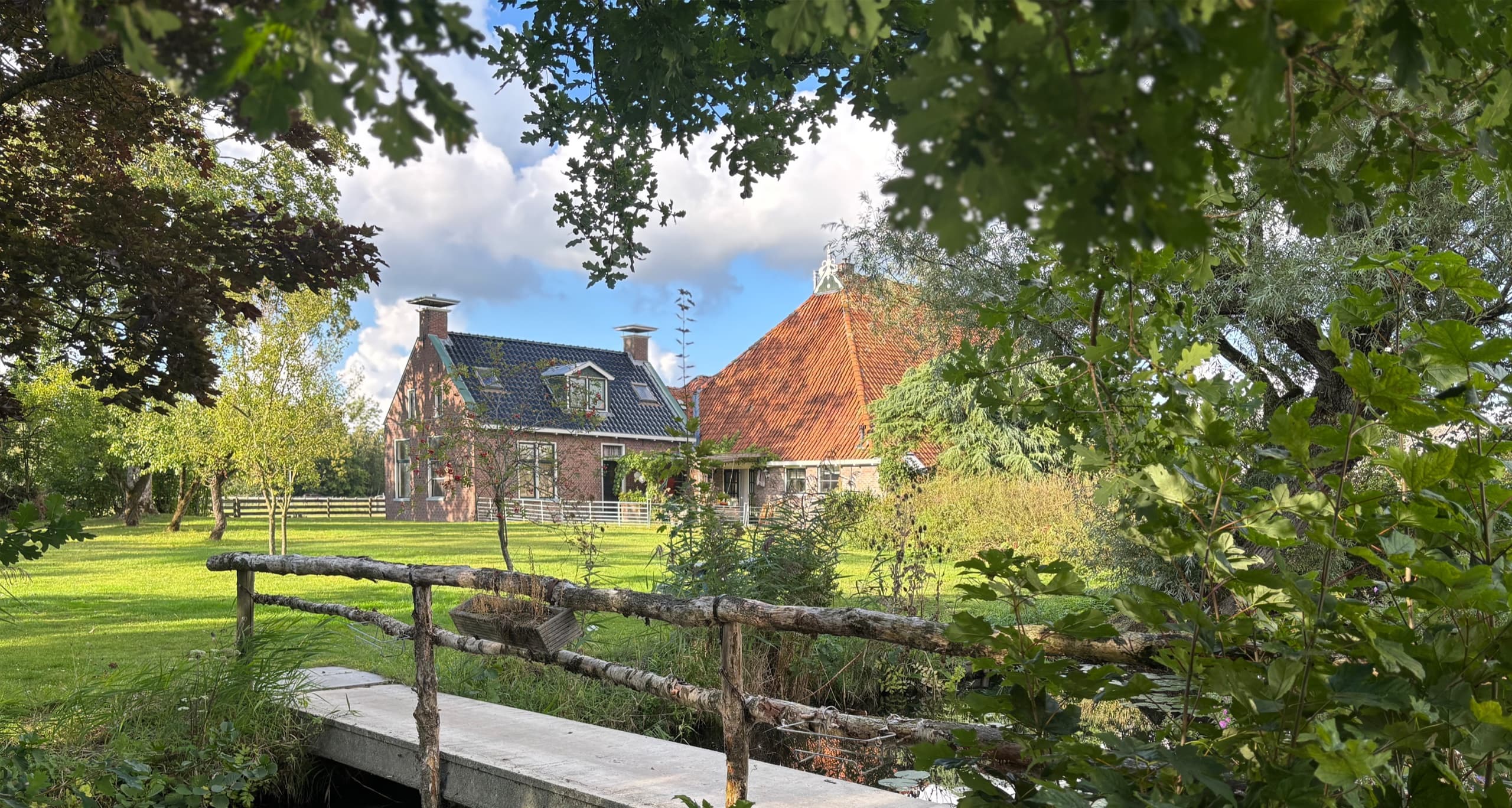 Uitzicht op de boerderij van Hiddema State in Hempens, gezien vanaf een bruggetje over de sloot, met een groen erf en bomen op de voorgrond.