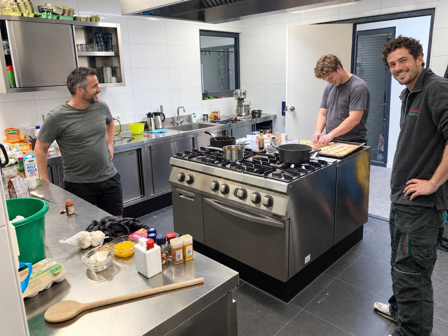 Drie mannen in Van Beets-kleding aan het werk in een professionele keuken met een groot gasfornuis. Deze foto is met AI bewerkt.