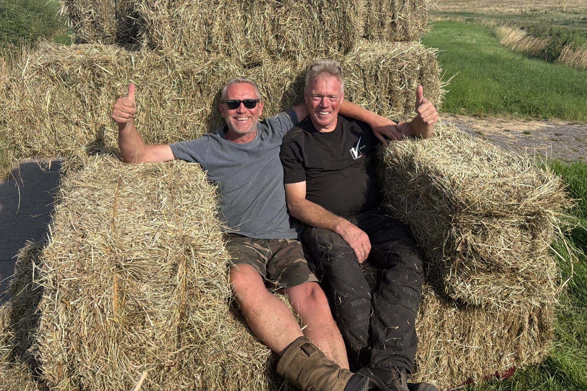 Twee collega's van Van Beets zitten breed lachend met opgestoken duimen op een berg hooibalen, op een erf met weilanden en een kerktoren in de verte.