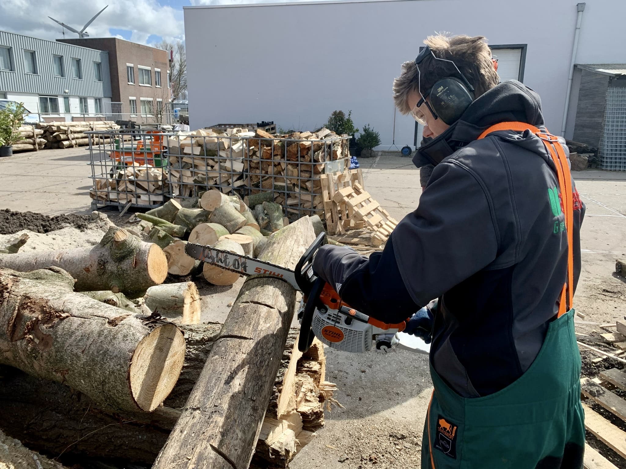 Een deelnemer met gehoorbescherming en veiligheidsbril zaagt met een kettingzaag een stam door op het werkerf van Van Beets.