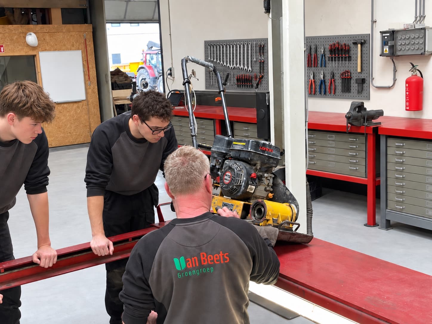 Twee jonge deelnemers en een begeleider in werkkleding van Van Beets Groengroep, gebogen over een trilplaat en een stalen profiel op de werkbank in de werkplaats aan de Uranusweg. Deze foto is met AI bewerkt.