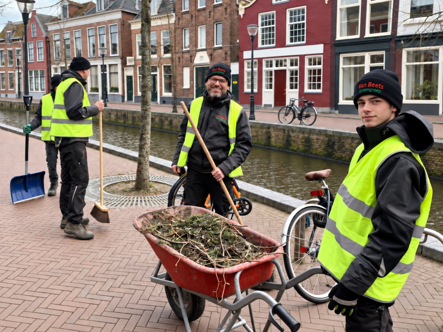 Een ploeg van Van Beets in veiligheidshesjes ruimt afgeknipt groen op langs een gracht in de Leeuwarder binnenstad, met een kruiwagen vol snoeiafval. Deze foto is met AI bewerkt.