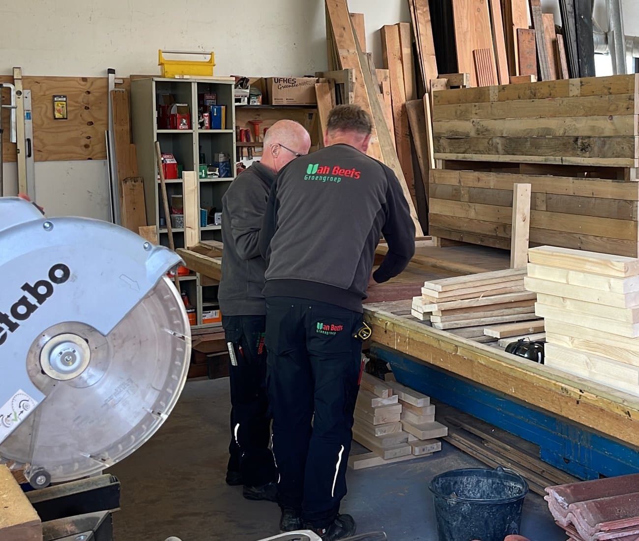 Twee collega's in werkkleding van Van Beets, gebogen over de werkbank in de houtwerkplaats, met een afkortzaag op de voorgrond.